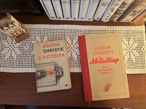 Two books over a table with a crochet runner, a miniature building and part of the spines of some books.