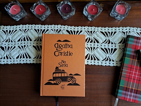 Um livro cor de salmão com uma carrinha preta desenhada e as palavras Agatha Christie Na Síria na capa, em cima de uma mesa de madeira escura, com um naperon de crochet branco, cinco castiçais com velas de vários tons de vermelho e um caderno com a capa forrada com tecido escocês em tons de vermelho e verde.