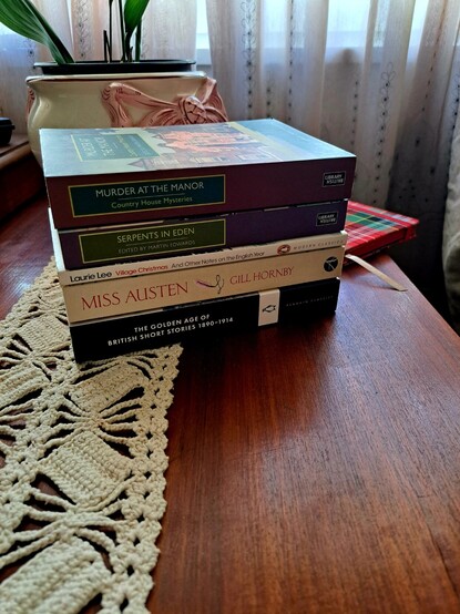 A pile of five books, on top of a wooden table, with a white and pink vase, part of a notebook, and curtains at the back.