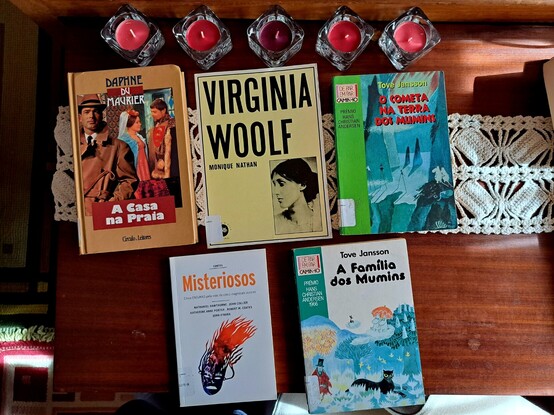Cinco livros em cima de uma mesa com um naperon branco de crochet e cinco velas vermelhas em castiçais baixos de vidro transparente
