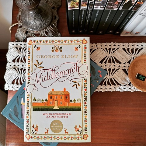 The book Middlemarch by George Eliot over a blue book both over a wooden table with a crochet doily. On the upper left part of a lamp and on the right spines of some black and white books. On the side a cork dish with a bookmark with the green ampelmännchen on.