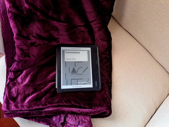 An ereader showing the cover of Project Gutenberg's book The Haunted Bookshop over a purple blanket over part of an armchair.