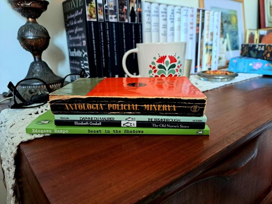 A stack of books over a wooden table with a tea cup, a lamp, eye  glasses and some other books I in the background