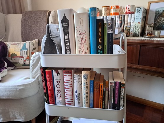 An armchair with a pillow depicting Tallinn next to a reading cart with my TBR for 2024. Behind, more books and cups on a wooden vanity.