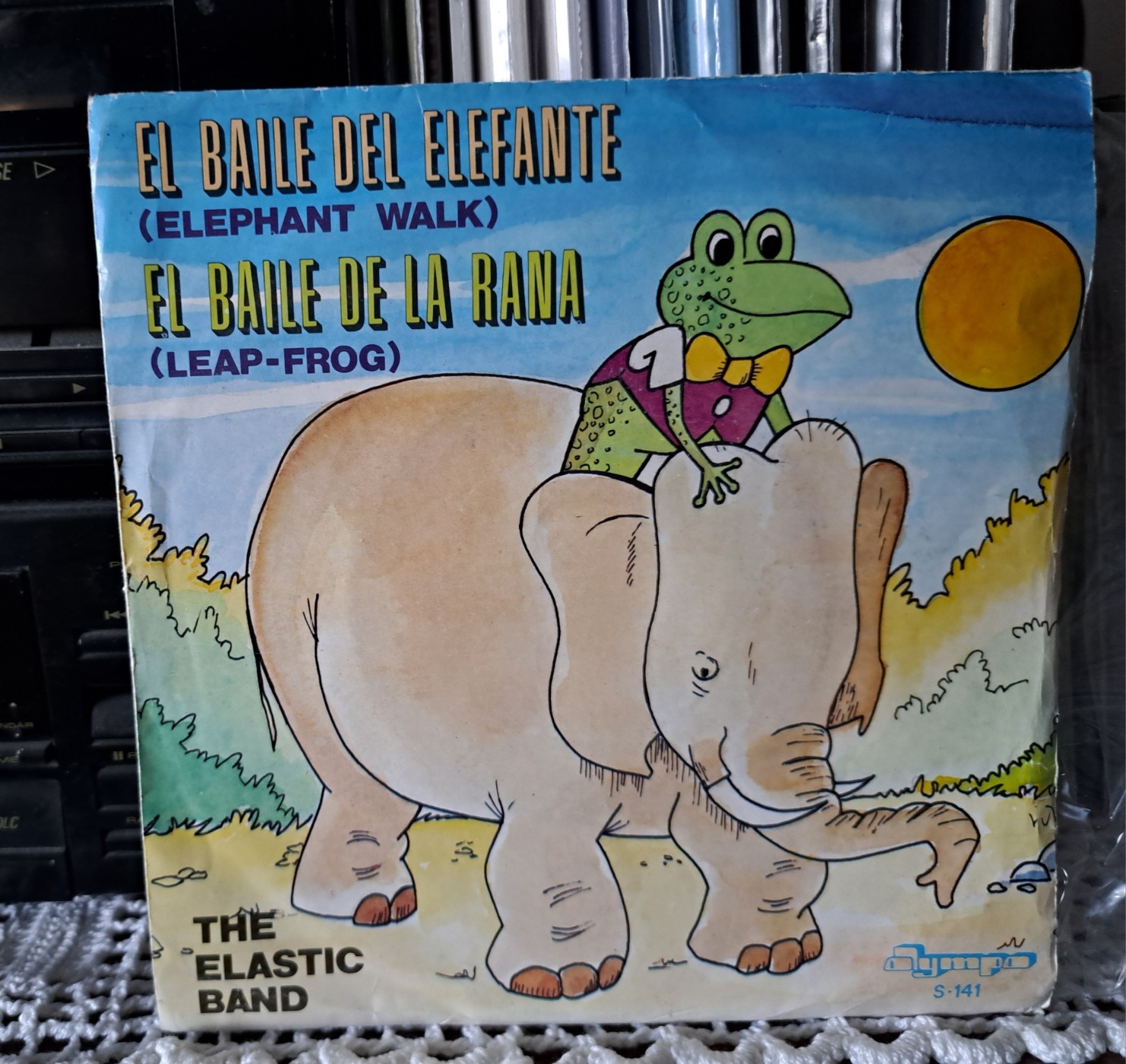 The Elastic Band: El Baile Del Elefante (Elephant Walk) El Baile De La Rana (Leap-Frog)  A 7" record sleeve, with the drawing of a frog 🐸 riding an elephant 🐘 under the sun 🌞