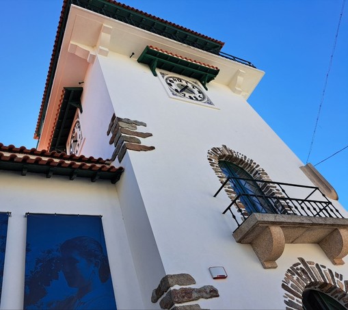 A white tower with wall clocks in each side seen from below with a balcony and some stones decorating the walls.