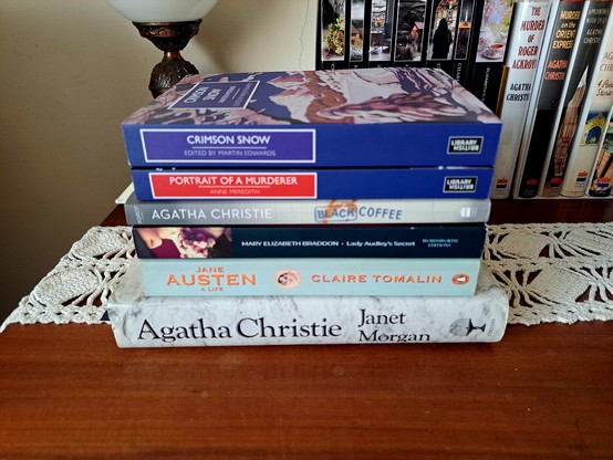 A pile of books in front of a table lamp and other books over a wooden table