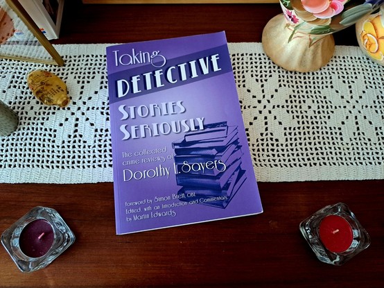 Um livro com capa roxa em cima de uma mesa de madeira. O livro reúne as críticas que Sayers publicava em jornais sobre os policiais que iam sendo publicados