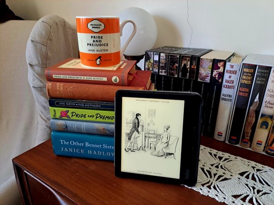 A stack of books with different editions of Pride and Prejudice, as well as other retellings, with an orange mug on top depicting the Penguin edition of the book and an ereader showing a drawing of a scene in the book. The books are on a wood table in front of other books and next to a (very comfy) armchair.