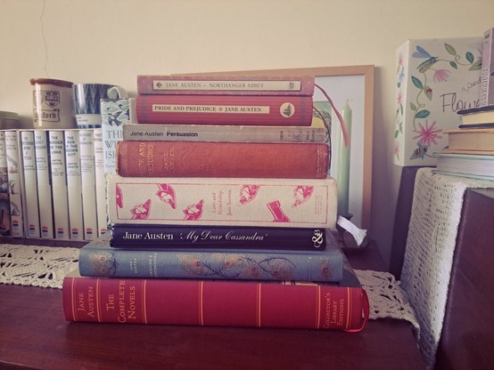 A stack of 8 books by Jane Austen on top of a wooden table in front of other books and two cups