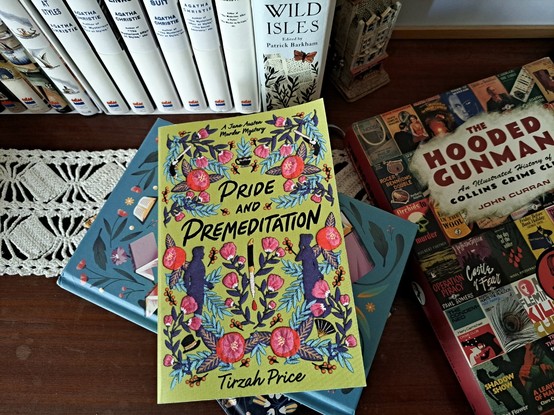 The book Pride and Premeditation, a green book with flowers and two person silhouette over a blue book in front of other books and next to another one in a wooden table