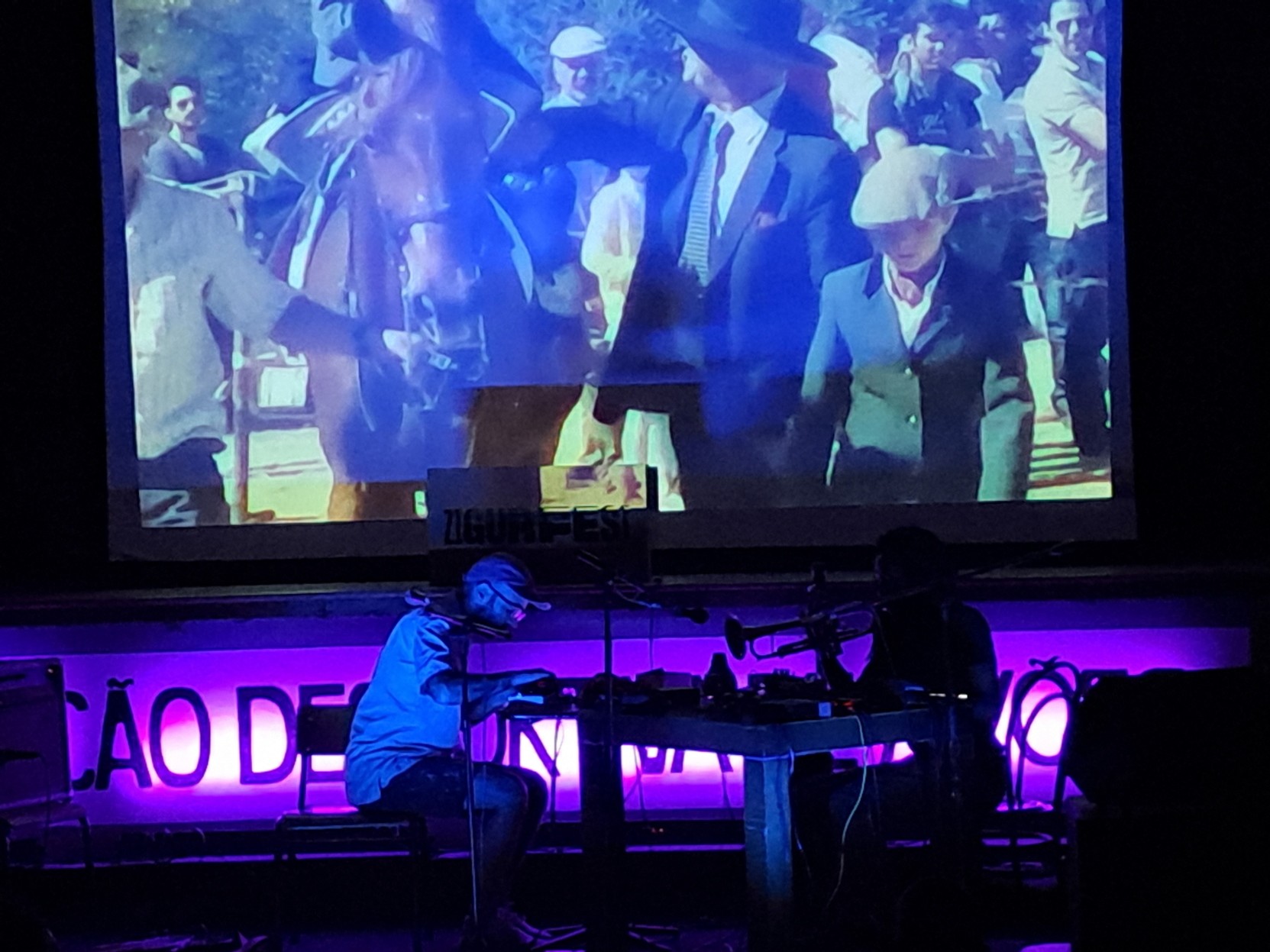 Sat on a table, Norberto Lobo on the left, playing on a keyboard, and Yaw Tembe on the right with a trumpet. Beyond them on the screen, a video from Lamego.