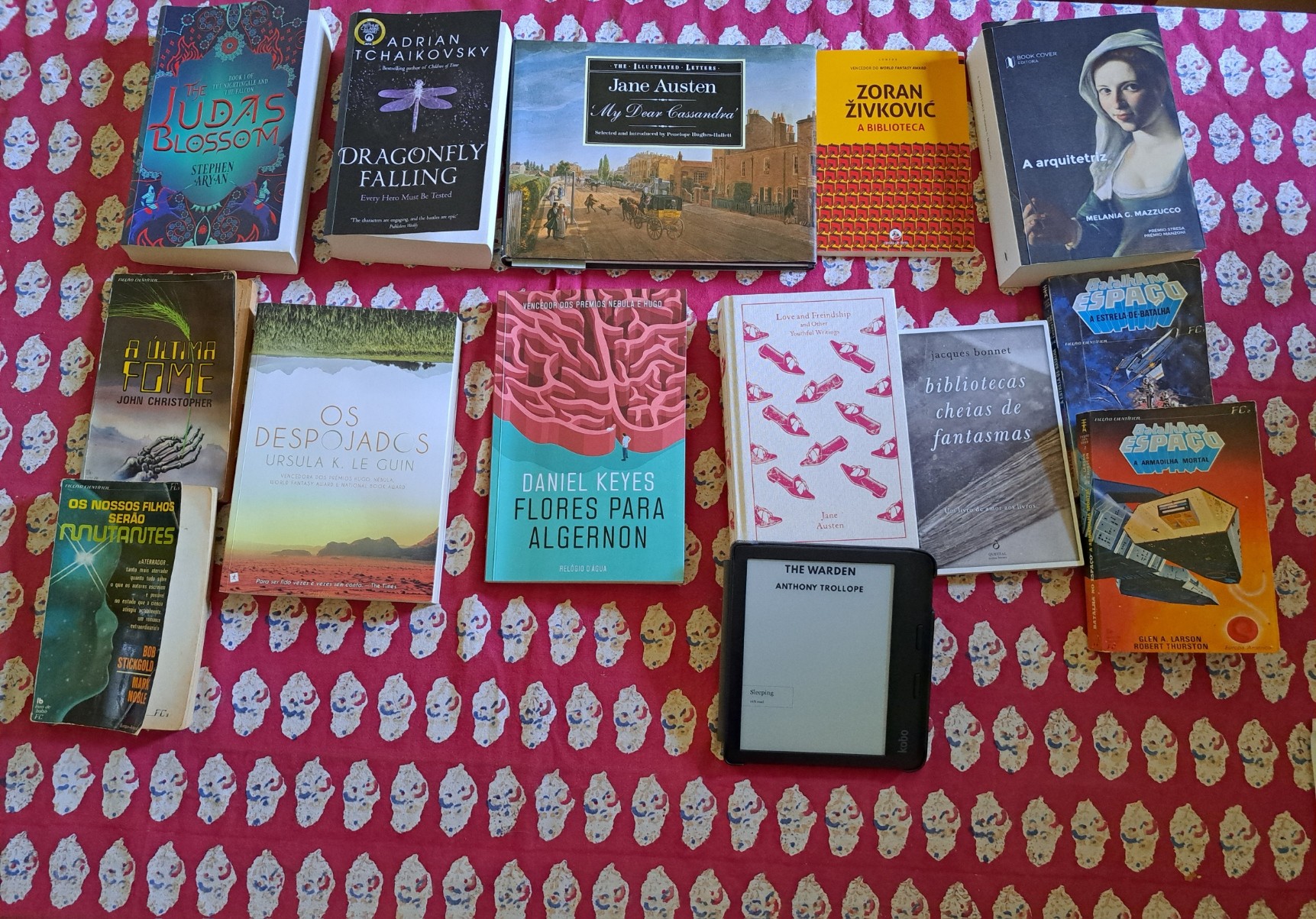 Uma mesa com 14 livros - ou 13 e um ebook reader, mas esse também está a mostrar e representar um livro.  De cima para baixo, esquerda para a direita: 1) The Judas Blossom 2) Dragonfly Falling 3) My Dear Cassandra 4) A Biblioteca  5) A arquitectriz 6) A última fome 7) Os nossos filhos serão mutantes  8) Os despojados  9) Flores para Algeron 10) Love and Freindship  11) The Warden 12) bibliotecas cheias de fantasmas 13) Batalha no Espaço: a Estrela-de-Batalha 14) Batalha no Espaço: a armadilha mortal