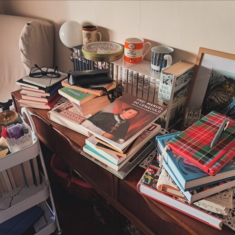 Piles of books, mugs, an ereader, and eyeglasses on a wood table with a lamp next to part of an armchair and a book cart