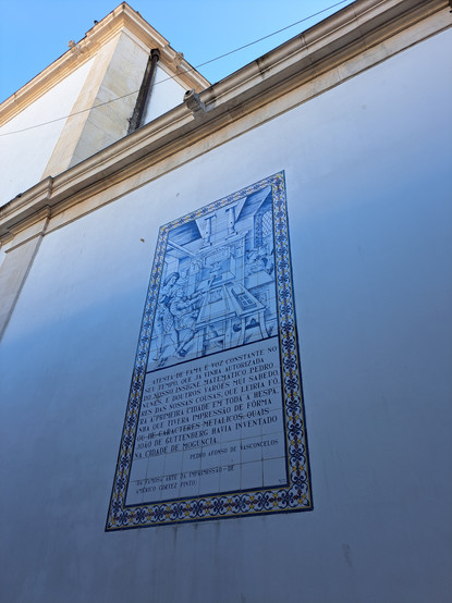 A church's outside wall with a blue and white tile panel depicting an old printing press