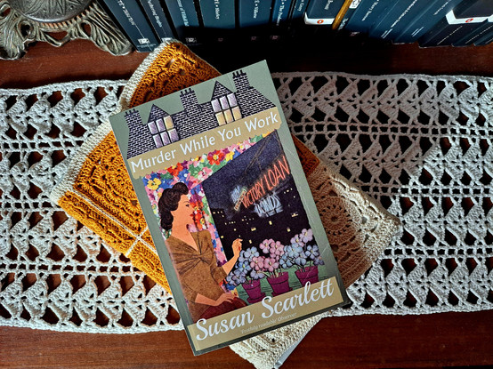 The book Murder while you work over another books with a crochet cover in front of several books and a lamp base on the top of a wooden table with a crochet doily 