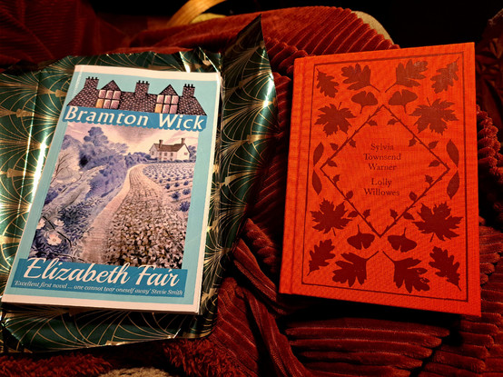 The books Bramton Wick and Lolly Willowes over a gift wrapping paper green and gold over a red blanket 