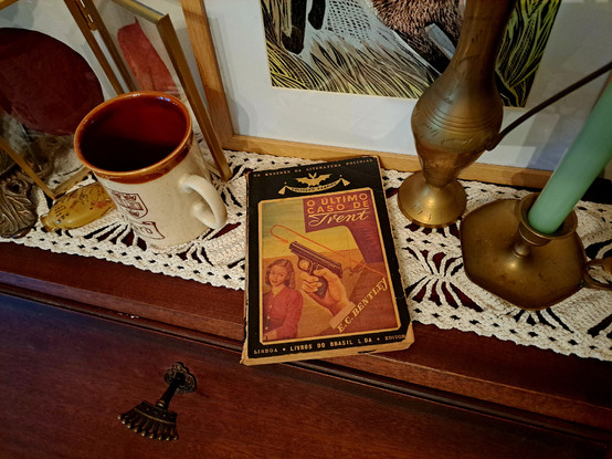 A book over a wooden closed desk with the key in the lock, showing a candle, a small metal vase, autumn leaves on a frame and a cup