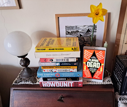 All the books by The Detection Club I have, on top of a wooden desk with an old lamp on one side and on the other a tablet showing the red cover of the most recent book, standing in front of a jar with a yellow daffodil