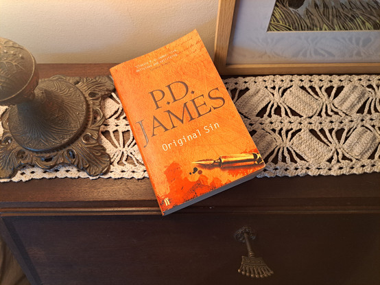 A book with an orange cover (Original sin by P. D. James) over a writing desk with a key in the lock a base of lamp with the light on over the book