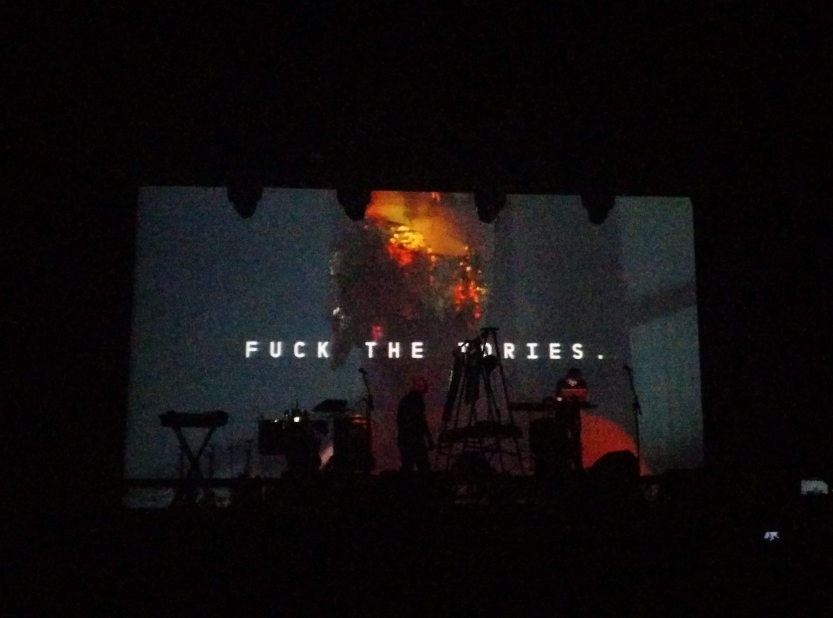 Test Dept. no palco. No ecrã atrás deles a mensagem é &ldquo;Fuck the Tories.&rdquo;