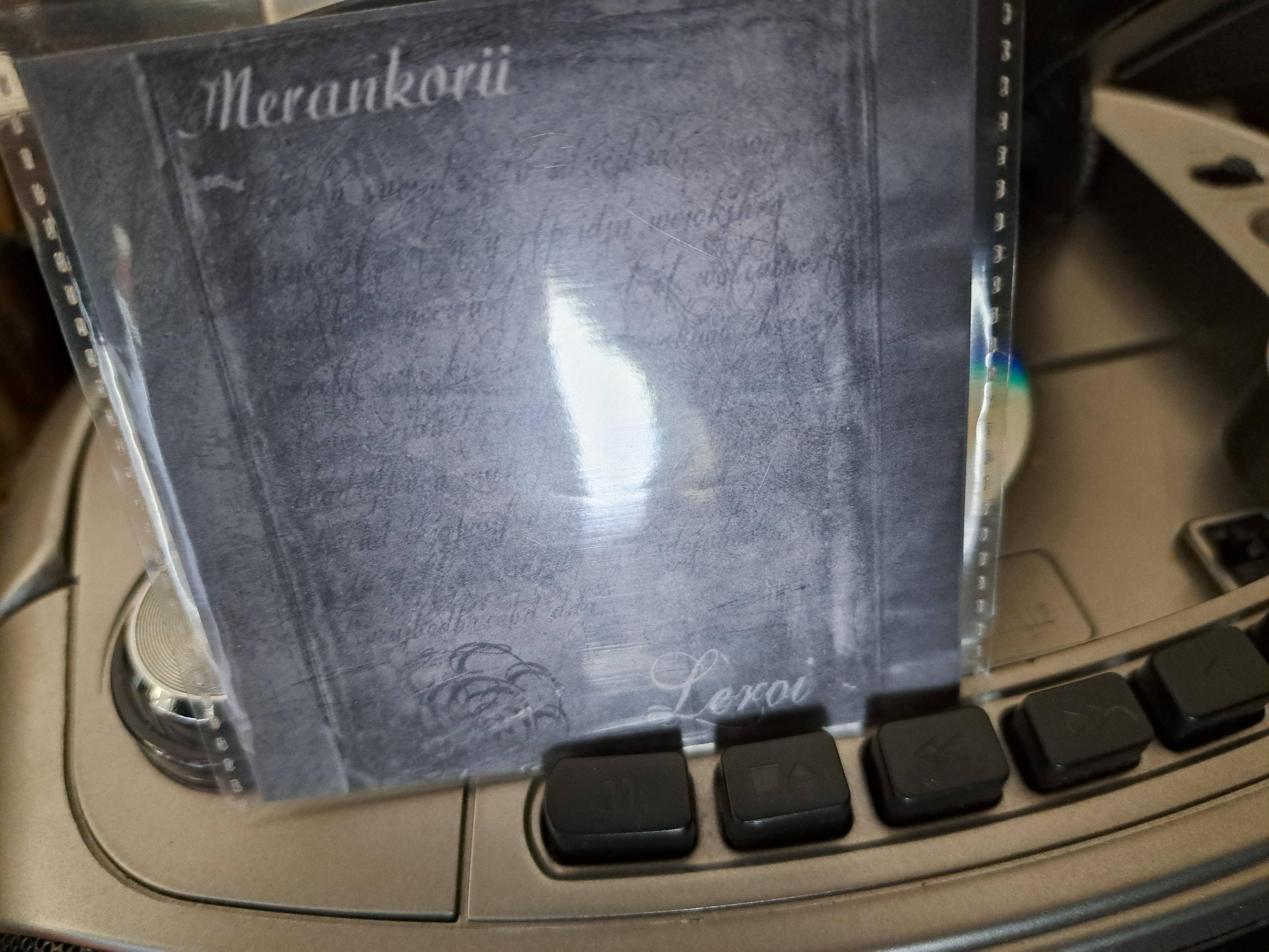 Foto do CD dentro de um leitor, e a capa do CD em frente. A capa é uma imagem a preto e branco de algo escriturado, dizendo no canto superior esquerdo "Merankorii" e no canto inferior direito "Leroi".